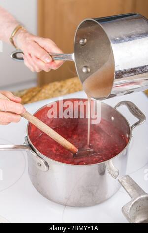 Donna versando di pectina di frutta e di acqua nella miscela di lamponi come una parte di confettura di lamponi. (MR) (PR) Foto Stock