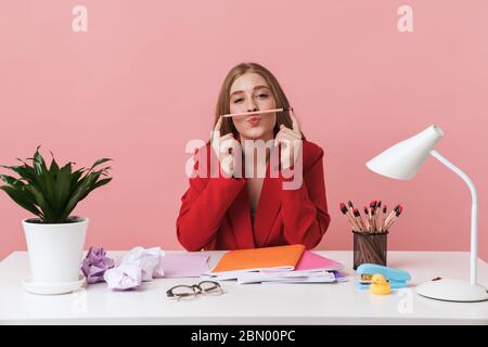 Immagine di una giovane donna carina divertente sedersi al tavolo al chiuso isolato su sfondo rosa parete tenendo matita. Foto Stock