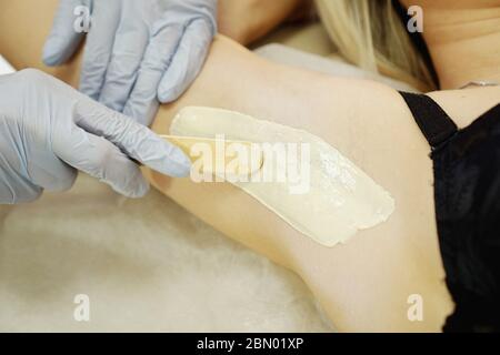 Ascella donna cerata con spatola. Procedura di epilazione con cera da salone. Ceratura corpo femminile per la rimozione dei capelli da parte del terapista primo piano Foto Stock