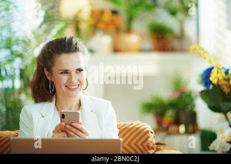 happy housewordie trendy 40 anni in blusa bianca e giacca con computer portatile e smartphone utilizzando applicazioni a casa moderna in giorno di sole. Foto Stock