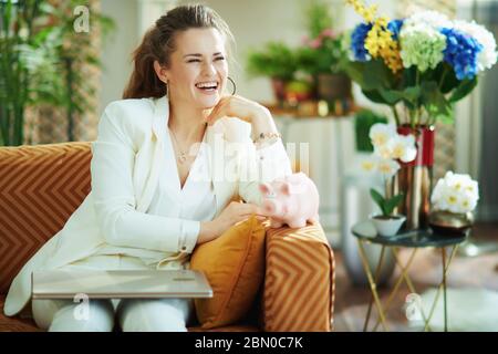 sorridente elegante casalinga di mezza età in blusa bianca e giacca con computer portatile e pig bank guardando in lontananza nel moderno soggiorno di sunn Foto Stock