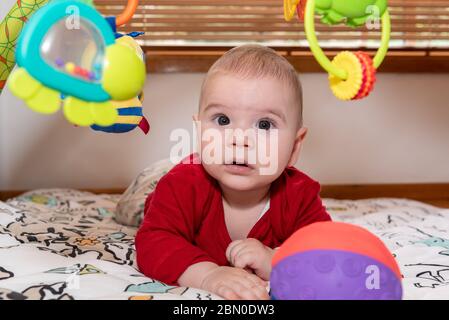 Bambino carino durante il pancino guardando la macchina fotografica. bambino di 6 mesi con espressione di curiosità sul suo viso circondato da giocattoli colorati. Foto Stock