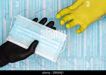Il medico tiene la maschera chirurgica per il viso in mano nel guanto medico nero e la dona al paziente in guanto protettivo giallo. Concetto di medicinale sullo sfondo di Foto Stock