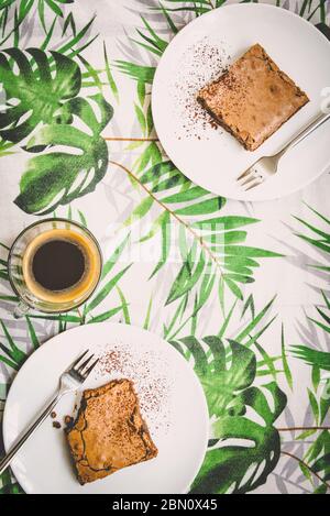 Vista dall'alto di una tazza di caffè e due piatti da dessert con torta di brownie su un panno da tavolo con motivo a foglie verdi. Foto Stock