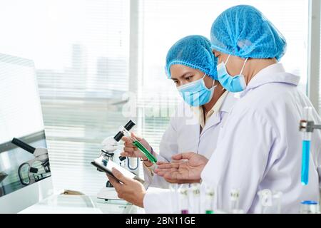 Operatori di laboratorio seri con provetta di liquido verde che discute i risultati della ricerca e li confronta con le informazioni sullo smartphone Foto Stock