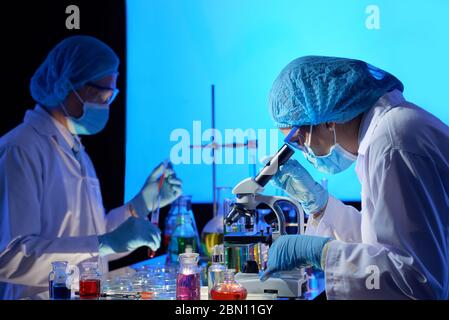 Team di scienziati che lavorano sul vaccino covid-19 in laboratorio scuro, mescolando liquidi e guardando attraverso il microscopio Foto Stock