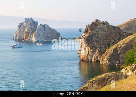 La costa rocciosa dell'isola di Olkhon e di Capo Burhan in una serata estiva ai raggi del sole che tramonta. Barche da diporto nelle acque blu del lago Baikal Foto Stock