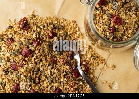 Granola fatta in casa con semi e mirtilli secchi su una carta da forno. Sana colazione vegana, spuntino o pranzo. Vasetto di vetro per limatura con cucchiaio di metallo. Vista dall'alto. Foto Stock