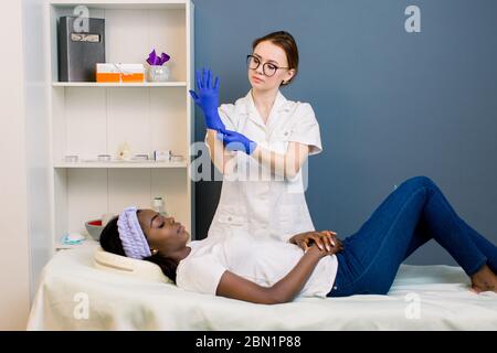 Cosmetologia professionale. Donna positiva molto felice, cosmetologa in piedi vicino al suo cliente, ragazza africana che si trova sul tavolo della spa mentre indossa blu Foto Stock