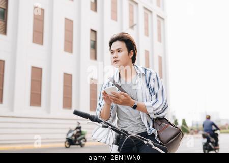 Elegante uomo chattare mentre si utilizza il telefono cellulare mentre è seduto sulla bicicletta, all'esterno. Vestito in plaid shirt, t-shirt e jeans. Close-up. Foto Stock