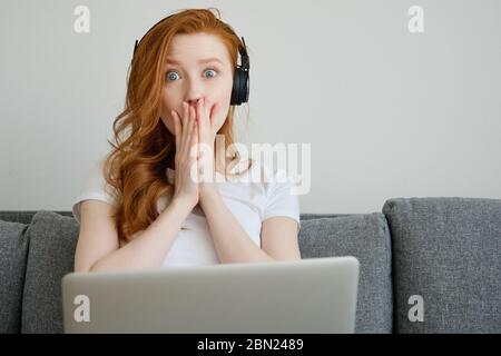 Ragazza rossa in una T-shirt e cuffie si siede su un divano con un computer portatile e copre la bocca con le mani, gli occhi larghi, guarda nella cornice Foto Stock