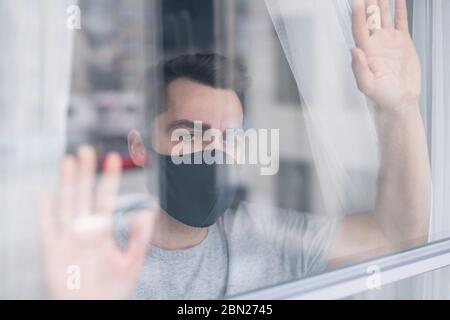 Dietro la vista di vetro di triste giovane uomo in maschera nera protettiva tenendo le mani sulla finestra e contemplando il paesaggio esterno, vuole uscire di casa Foto Stock