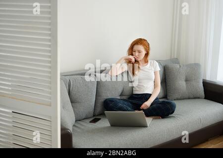 Una ragazza dai capelli rossi nei vestiti domestici si siede appoggiata su un divano e guarda pensierosamente in un laptop davanti a lei. Foto Stock