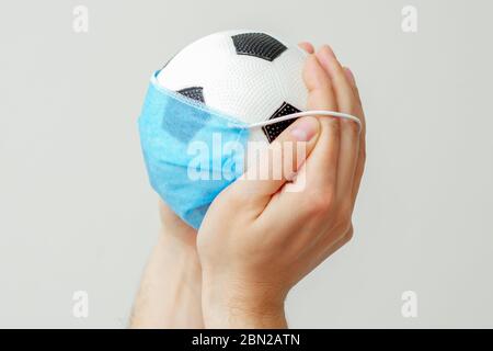 Primo piano di palla di calcio con una maschera medica nelle mani dell'uomo su uno sfondo chiaro. Cancellazione di eventi sportivi. Foto Stock