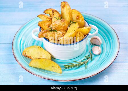 Patate fritte incastrano le patate in un piatto blu su sfondo di legno blu con aglio e un ramo di razmarin. Foto Stock