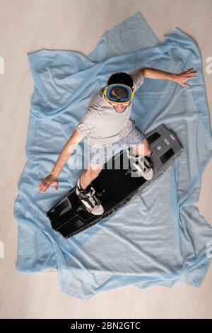 Vista sopra di giovane felice in homewear e maschera bilanciamento con braccia estese su snowboard a casa isolamento Foto Stock