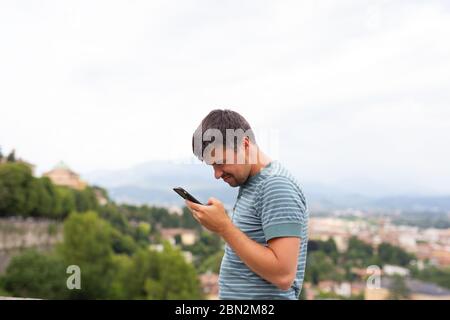 Giovane uomo orologi surf internet tiene sguardi smartphone cellulare in una città. Concetto di viaggio. Viaggiatore. Vacanze estive. Fine della quarantena terminata Foto Stock