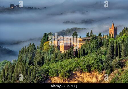 Il campanile dell'Abbazia di Monte Oliveto è immerso in una fitta nebbia autunnale nel paesaggio delle crete senesi vicino a Siena Foto Stock