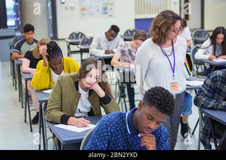 Insegnante di scuola superiore che supervisiona gli studenti che prendono banchi di esame in classe Foto Stock