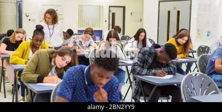 Insegnante di scuola superiore che supervisiona gli studenti che si stanno prendendo in esame alle scrivanie Foto Stock