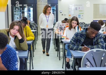 Insegnante di scuola superiore che supervisiona gli studenti che si stanno prendendo in esame alle scrivanie Foto Stock