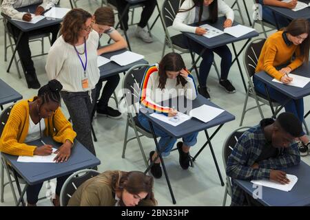 Insegnante di scuola superiore che supervisiona gli studenti che si stanno prendendo in esame ai tavoli Foto Stock