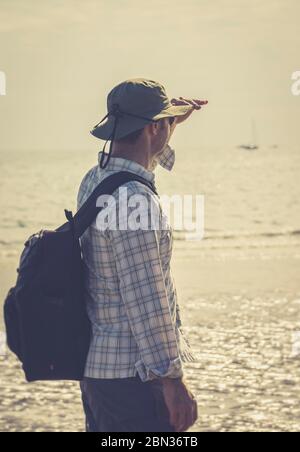 Vista posteriore da vicino di uomo isolato britannico villeggiante indossare cappello da sole sulla spiaggia britannica guardando verso il mare. Vacanza estiva staycation, uomo scherma occhi. Foto Stock