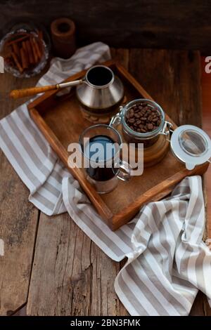 Una tazza di caffè su un vassoio di legno Turk e chicchi di caffè. Fai colazione a casa. Ancora vita con una bevanda in un bicchiere con Cezve. Tovaglia a righe su Foto Stock