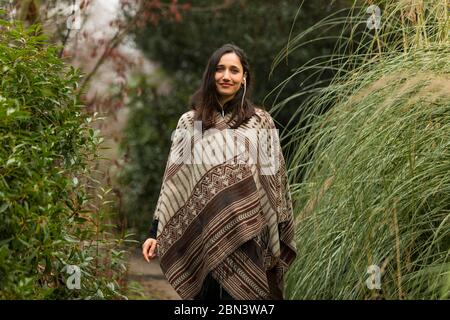 Bella donna indiana che indossa poncho in posa tra fogliame autunnale e sorridente alla macchina fotografica. Foto Stock