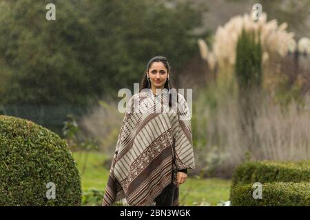 Vista frontale ritratto di donna indiana che indossa poncho in giardino ornamentale. Foto Stock