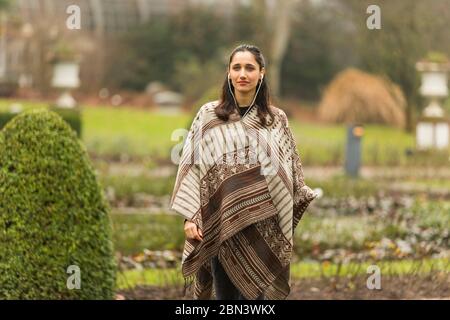 Donna indiana che indossa poncho e ascoltare gli auricolari in piedi in giardino ornamentale topiario. Foto Stock