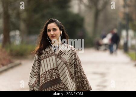 Giovane donna indiana che indossa un poncho e ascolta gli auricolari che posano sulla strada del parco. Foto Stock