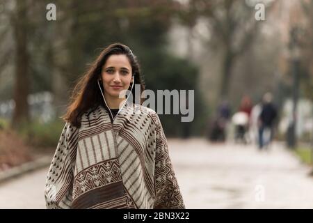 Giovane donna indiana che indossa poncho e auricolari sorridenti per la macchina fotografica all'aperto. Scatto medio. Foto Stock