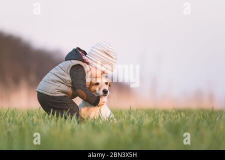 Piccolo bambino in giacca gialla con jack russel terrier cucciolo abbracciarsi l'un l'altro sul campo di primavera al tramonto Foto Stock