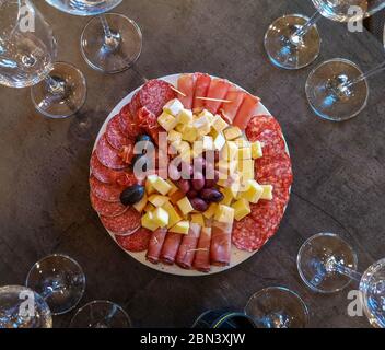Vista dall'alto di un tavolo con bicchieri vuoti e un piatto con vari salumi: Prosciutto a fette, pancetta, salame, cubetti di formaggio, olive rosse e nere, servito come st Foto Stock