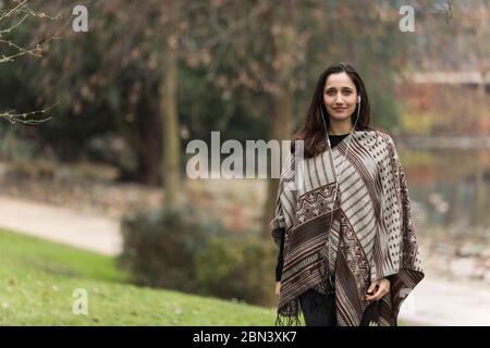 Giovane donna medio orientale che indossa poncho a motivi geometrici e ascolta le cuffie nel parco. Foto Stock