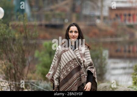 Scatto medio di modello femminile dai capelli scuri che posa in poncho all'aperto. Vista frontale. Foto Stock