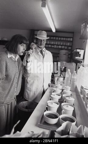 1980s, degustazione di tè..... Un uomo con un cappotto bianco e un cappello, un assaggiatore esperto, in piedi accanto a una donna, entrambi usando cucchiai per assaggiare il tè, Harrogate, Inghilterra, Regno Unito. Il tè, la pianta Camelia Sinensis, ha sapori e aspetto diversi dalle condizioni di coltivazione e dai processi di produzione. Foto Stock