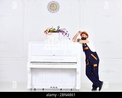 Bel lavoratore che tiene il suo casco protettivo. Uomo stanco che si gode una breve pausa. Pianoforte bianco vintage con tastiera aperta e vaso in vetro con fiori rosa e viola isolati su sfondo bianco. Foto Stock