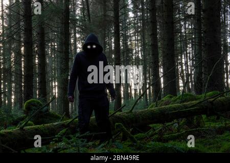 Un concetto di orrore di una foresta invernale spaventosa con una figura spaventosa con cappuccio e occhi scintillanti accanto a un albero caduto Foto Stock