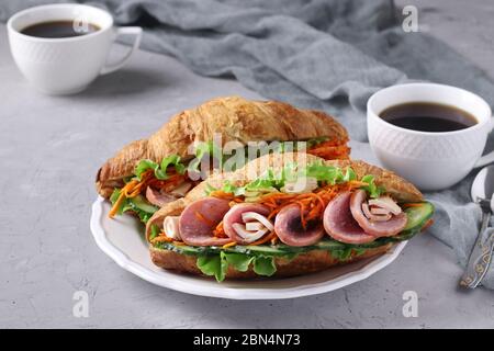 Due croissant-sandwich con lattuga, carote, cetrioli, salsiccia, formaggio e due tazze di caffè su fondo grigio chiaro in cemento. Colazione o. Foto Stock