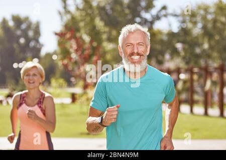 Essere in forma e in salute. Felice uomo maturo sorridente alla macchina fotografica mentre corre insieme con sua moglie la mattina presto. Messa a fuoco selettiva Foto Stock