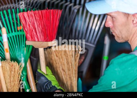Primo piano di Caucasian maschio Groundskeeper scelta piccola scopata in stoccaggio Shed. Foto Stock
