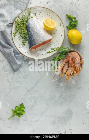 Trota circondata di prezzemolo, limone, gamberi, gamberi in piatto di ceramica. Superficie del tavolo in cemento grigio chiaro. Sano sfondo di pesce. Foto Stock