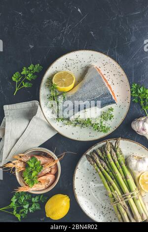 Trota circondata da prezzemolo, limone, gamberi, gamberi, asparagi in piatti di ceramica. Superficie del tavolo in cemento nero. Sano sfondo di pesce. Foto Stock