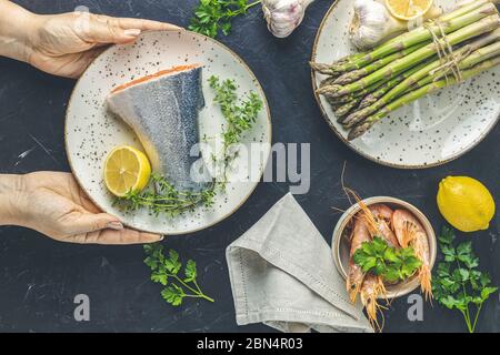 Donna tiene piatto in ceramica con trota cruda pesce, timo e limone in mano su piano di tavolo in cemento nero piastre circondate con asparagi crudi freschi, sh Foto Stock