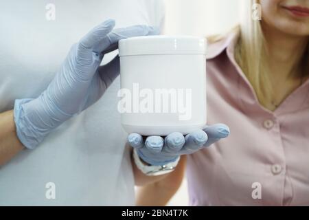 Il medico mostra il rimedio del paziente nel salone di bellezza. Cosmetologia il medico consiglia il paziente. Foto ravvicinata del vaso bianco con un cosmetico Foto Stock