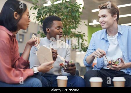 Gruppo multietnico di giovani che mangiano cibo da asporto e sorridono durante la pausa pranzo a scuola o in ufficio
