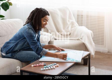 Quarantena Hobby. Rilassato Black Woman disegno pittura da numeri a casa Foto Stock