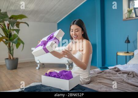 Giovane brunetta in abito da notte seduto a letto, sorridente, aprendo la scatola presente con nastro viola, guardando sorpreso Foto Stock